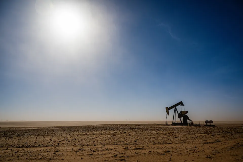 Distant view of oil derrick on barren plain with harsh sun in background.